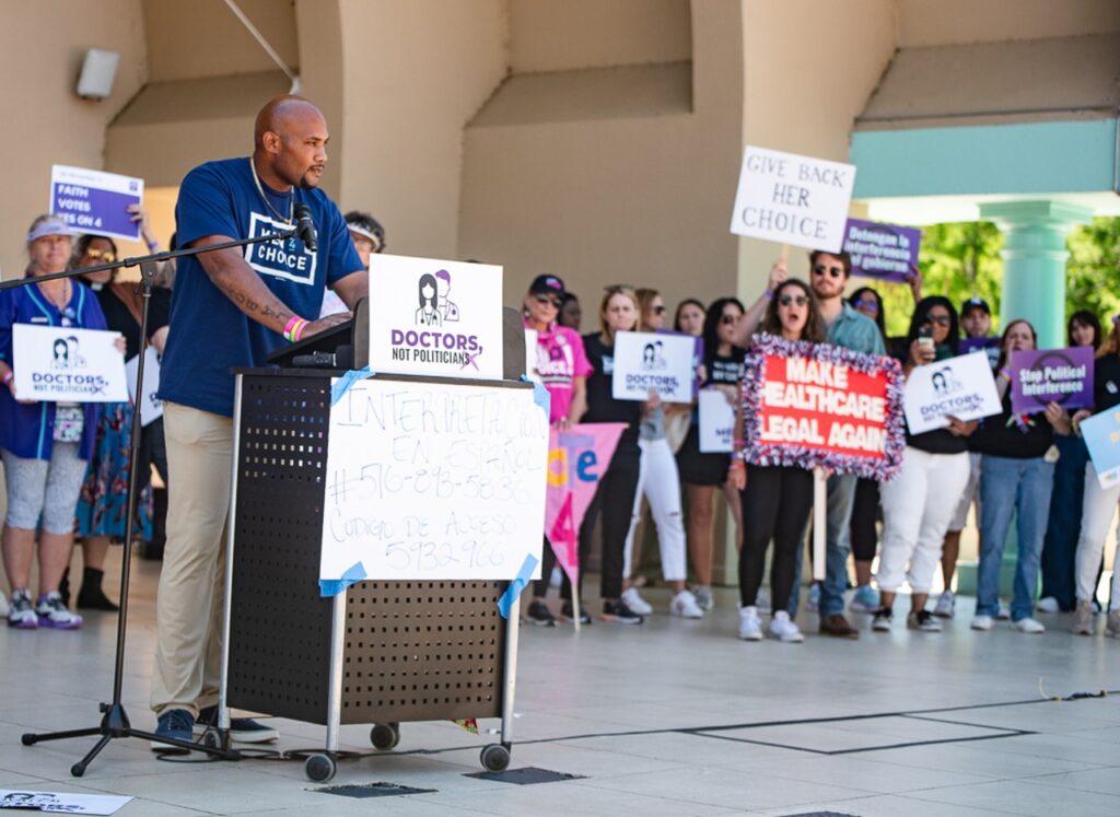 A Black man in a "Men 4 Choice" shirt stands at a podium which has a sign that says "Doctors not politicians". With him is a crowd of supporters to vote yes on Amendment 4 and end Florida's abortion ban.