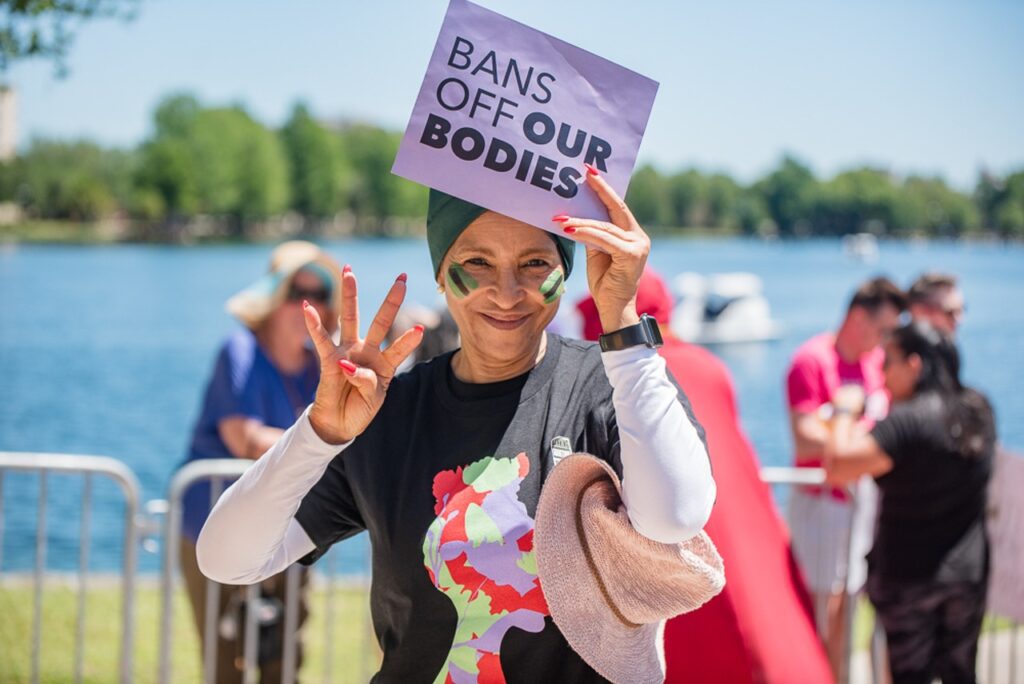 A Black woman wearing a black shirt with white sleeves holds up 4 fingers for Amendment 4 and a sign that reads "Bans off our bodies"